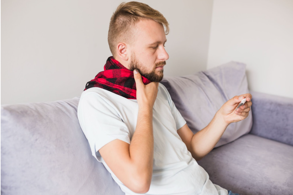 Мъж с шал и болки в гърлото държи термометър