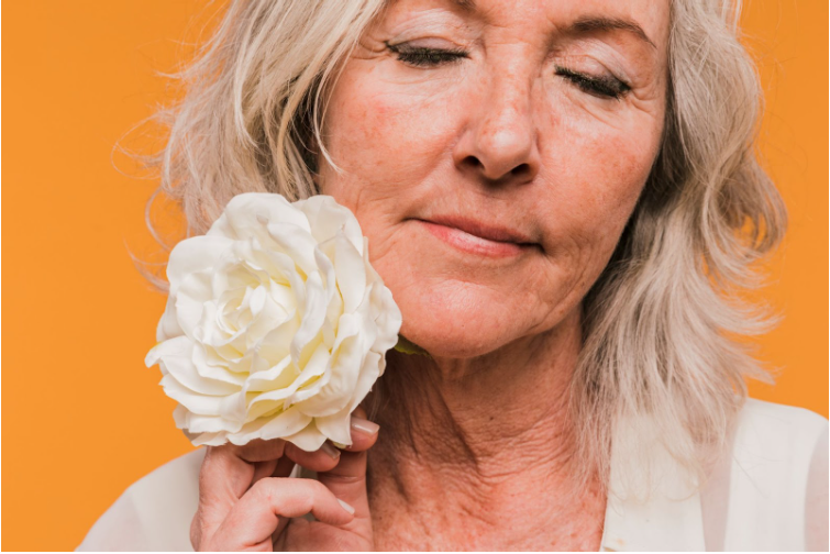 Възрастна жена с бръчки по лицето държи до лицето си бяло цвете